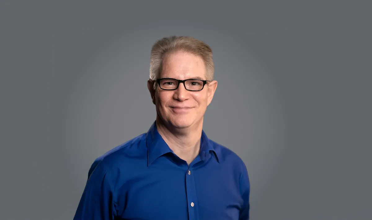 A smiling middle-aged Caucasian man with a flat-top haircut, glasses, and blue button-down shirt poses for the camera