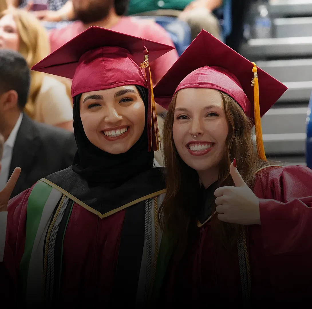 Two women wearing graduation gowns smiling and throwing a peace sign and a thumbs-up to the camera