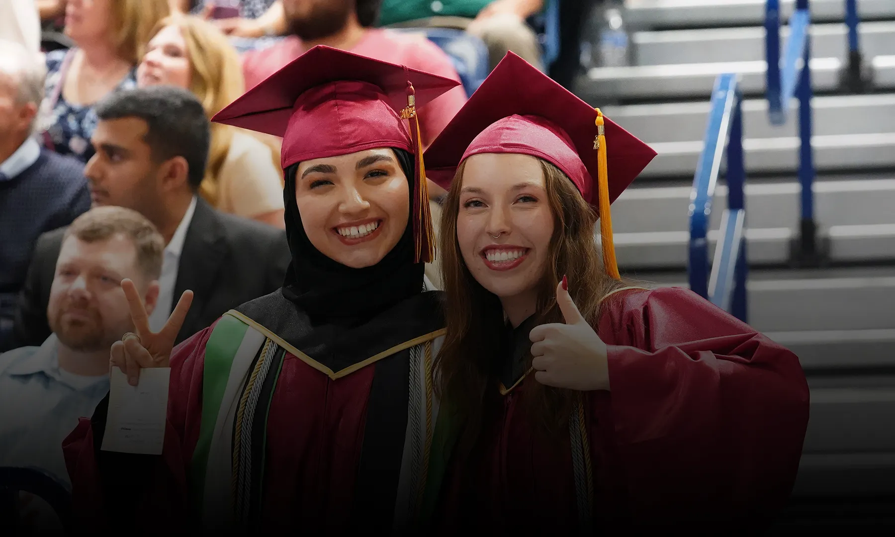 Two female students in graduation gowns smiling and throwing a thumbs-up and peace sign to the camera