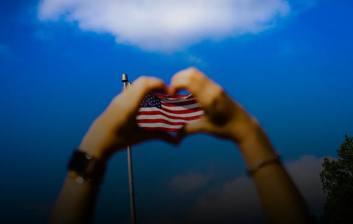 A person makes heart hands over an American flag flying in the background