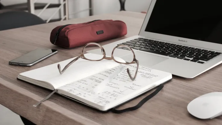 Desk with notebook, computer, glasses and phone courtesy of Dan Dimmock from unsplash.com