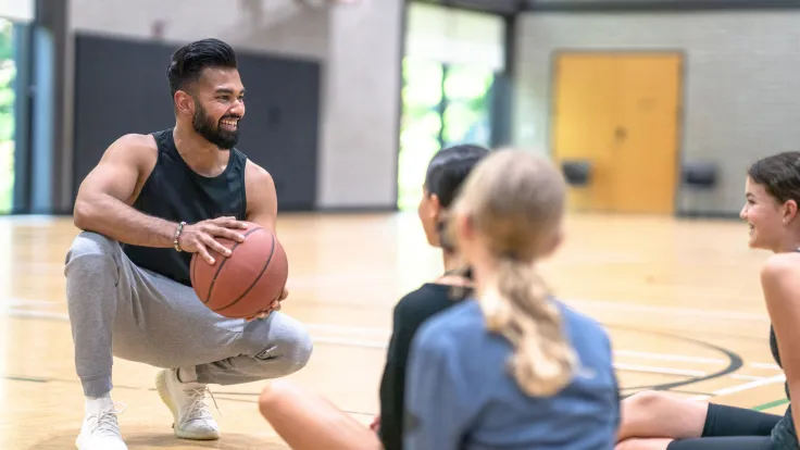 A male Physical Education teacher of Indian decent squats down in front of a small group of female students