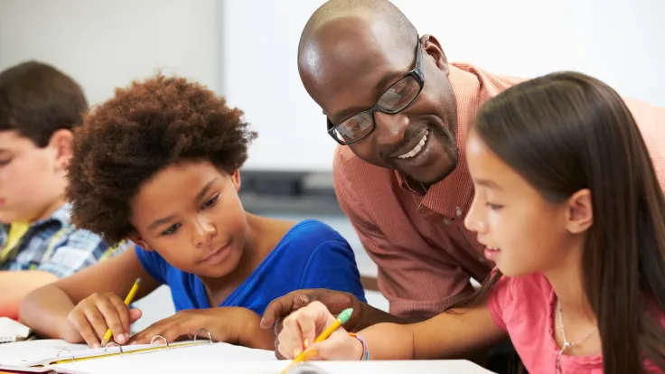 Teacher Helping Pupils Studying At Desks In Classroom Checking Work Smiling