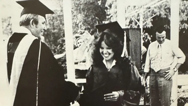 A sepia-toned historical photo of a smiling woman receiving her diploma at a commencement ceremony
