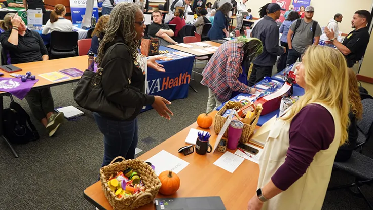 A woman stops by a table at a career fair to ask a vendor questions about their employment opportunities