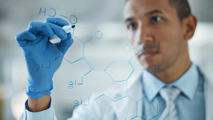 A chemical engineer in a lab coat and nitrile gloves writes a chemical formula on a board