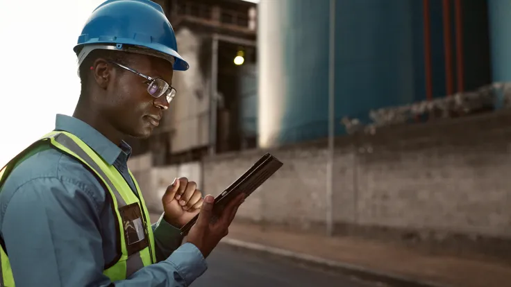 A civil engineer wearing a hard hat reviews information on a tablet at a job site