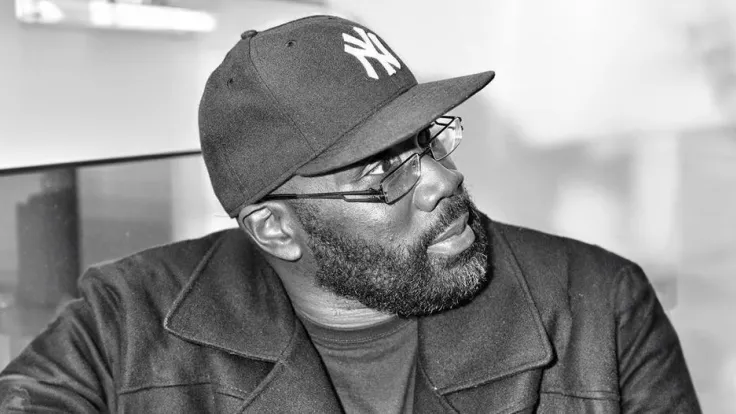 Black and white photo of Derek Tobias, a Black American man with glasses and a bear wearing a New York Yankees cap and a pea coat