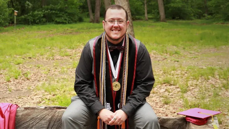 Jessie Harper sits smiling on a stone wall with graduation cap and gown settled down near him